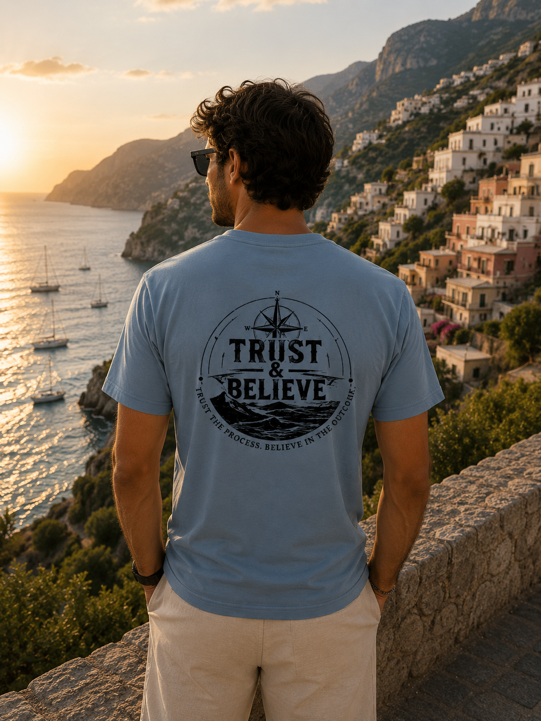Man wearing a 'Trust & Believe' t-shirt overlooking a scenic coastal town at sunset.