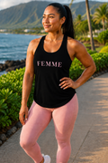 A woman wearing a black racerback tank top with 'FEMME' printed in pink across the chest, paired with pink leggings, standing outdoors on a path near the ocean with mountains and palm trees in the background.