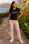 A woman stands outdoors on a stone path wearing a black short-sleeve T-shirt with the word 'FEMME' in white across the chest, light pink pants, and white sneakers. The background shows tropical greenery, flowers, water, and mountains.