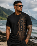A man wearing a black t-shirt with a brown tribal-style graphic print featuring the word 'MANAVA' vertically, standing on a beach with mountains and ocean in the background.